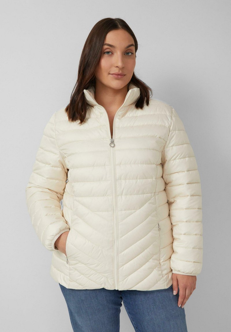Cremfarbener, wärmender Steppjacke mit hohem Kragen, mit einem gesteppten Design und seitlichen Taschen, getragen mit blauen Jeans. Glatte Textur.