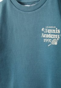 Zils kokvilnas džemperis ar ribotā apkakli un baltiem uzdrukātiem burtiem "Los Angeles Tennis Academy 1997" ar tenisa raketi grafiku.