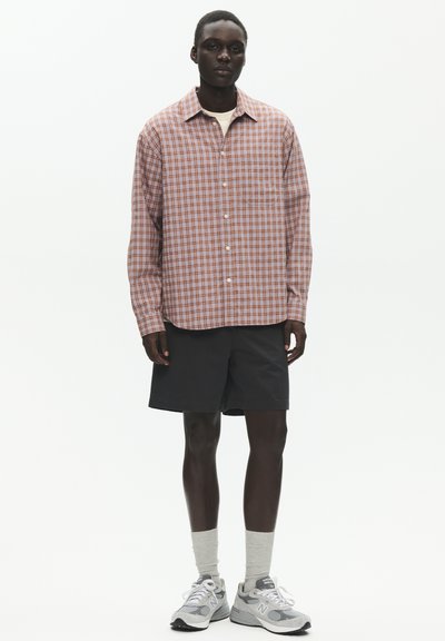 Homme portant une chemise à carreaux rouge et blanc surdimensionnée boutonnée, un short noir, des chaussettes blanches et des baskets New Balance grises, debout devant un fond blanc.