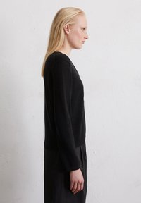 Schwarzer strukturierten Pullover mit langen Ärmeln und rundem Halsausschnitt, kombiniert mit einem schwarzen Rock, seitlich gesehen vor einem neutralen Hintergrund.