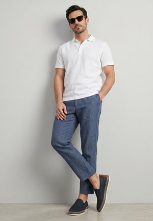 Man met zwarte zonnebril, wit poloshirt met korte mouwen, blauwe broek en marineblauwe instappers, leunend tegen een lichtgrijze muur met één hand in zijn zak.