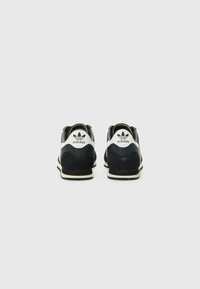 Baskets athlétiques en cuir et daim noir avec une semelle intermédiaire blanche et une semelle extérieure noire. Elles possèdent un talon noir et blanc avec le logo Adidas.