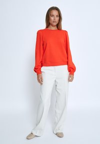 Orange sweatshirt with long, puffed sleeves, crew neckline, and a relaxed fit, paired with white tailored trousers and beige slip-on shoes.