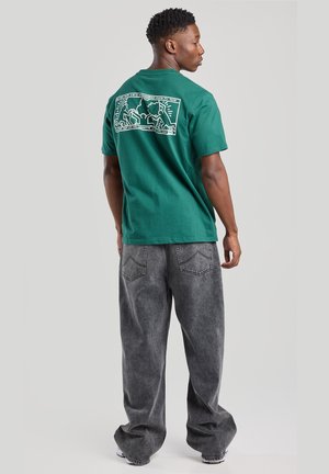 Green cotton t-shirt with a white graphic design on the back, paired with loose gray denim jeans featuring a faded texture and back pockets.