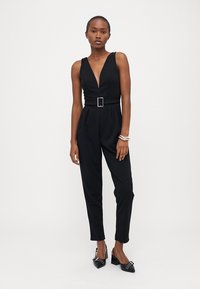 Μαύρο αμάνικο ολόσωμο φόρεμα με βαθύ V-ντεκολτέ, κεντημένη μέση με τετράγωνη πόρπη, στενές μπατζάκια και λεία υφή. Φορεμένο με μαύρες γόβες.