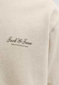 Bež teksturirana tkanina z majhnimi črnimi črkami, na katerih piše "Jack & Jones Archive Selection", natisnjenimi na površini.