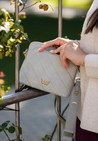 Světlé šedé crossbody taška vyrobená z texturovaného materiálu, s diagonálními švy, zlatým kováním a odnímatelným popruhem.