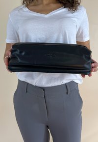 Bolsa de cuero negro con una textura suave y cierre de cremallera. Presenta un logo en relieve en la parte frontal y tiene forma rectangular.