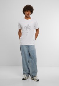Man met krullend haar, gekleed in een wit graphic t-shirt, lichtblauwe baggy jeans en zilveren sneakers, staande tegen een egale achtergrond.
