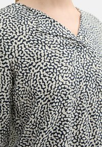 Marinebluse mit einem strukturierten Muster aus unregelmäßigen weißen Formen, einem umgeschlagenen Kragen und einem Knopfdetail am Ausschnitt.