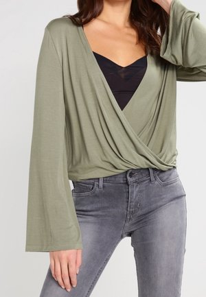 Kobieta w oliwkowozielonym długim rękawie typu wrap na czarnym topie na ramiączkach i szarych przecieranych dżinsach, pozująca z jedną ręką przy szyi.