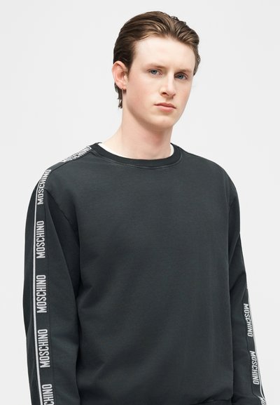 Νεαρός άνδρας με ανοιχτόχρωμο δέρμα και μαλλιά χτενισμένα προς τα πίσω, φορώντας μαύρο φούτερ Moschino με λωρίδες λογότυπου στα μανίκια, κοιτάζει μπροστά με ουδέτερη έκφραση.