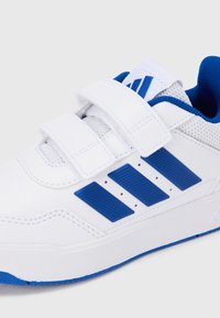 Zapatilla blanca con detalles en azul, que cuenta con dos tiras de Velcro, paneles de malla y tres franjas azules en el lateral. Textura sintética suave.