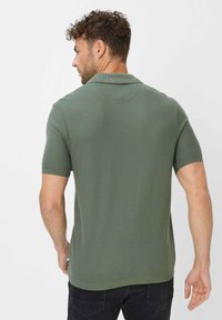 Strik polo skjorte i olivengrøn, med korte ærmer, krave og textured stof. Vist fra bagsiden med en diskret kantdetalje.