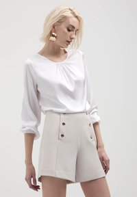 Blusa bianca a maniche lunghe con scollo arricciato abbinata a pantaloni corti beige a vita alta con dettagli in bottone dorato e tasche laterali.