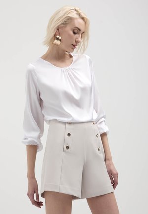 Blusa bianca a maniche lunghe con scollo arricciato abbinata a pantaloni corti beige a vita alta con dettagli in bottone dorato e tasche laterali.