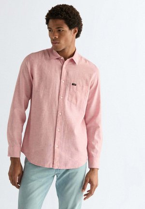 Mann trägt ein hellrosa Hemd mit Knöpfen und hellblaue Jeans, steht vor einem einfarbigen weißen Hintergrund.