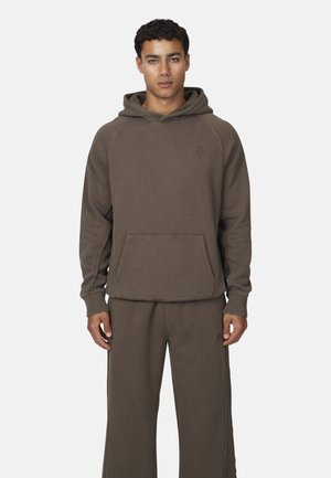 Homme portant un sweat à capuche marron et un pantalon large assorti, se tenant contre un fond clair uni.