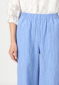 Ljusblå, strukturerade vida byxor med elastisk midja. Materialet verkar vara lätt och något skrynkligt, vilket ger en avslappnad passform.