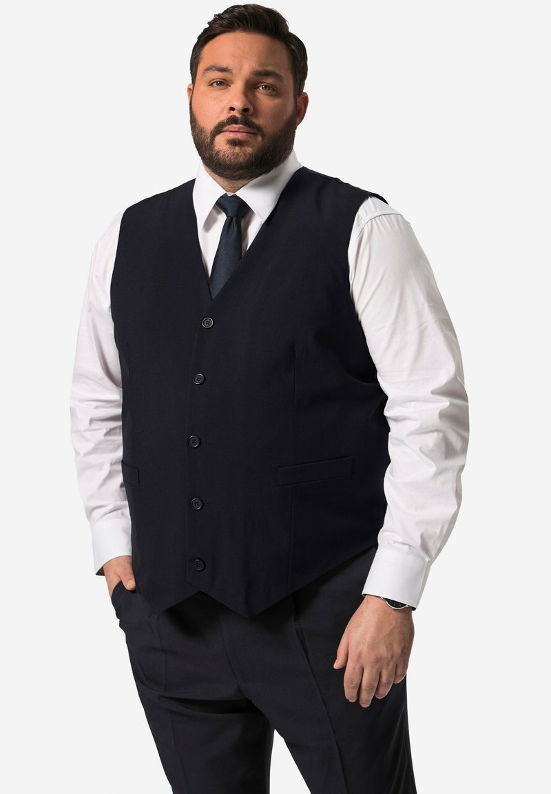 Gilet di un completo blu navy realizzato in un tessuto morbido, con sei bottoni e due tasche laterali, indossato sopra una camicia bianca e una cravatta scura, con una vestibilità sartoriale.