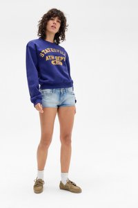 Sweatshirt roxo com "Statesville Ath Dept" em letras douradas, combinado com calções de ganga azul claro. Calçado castanho e meias brancas até ao tornozelo.