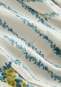 Tissu blanc doux avec des fines rayures florales bleues et une petite zone de fleurs jaunes, montrant la texture et les détails de la couture.