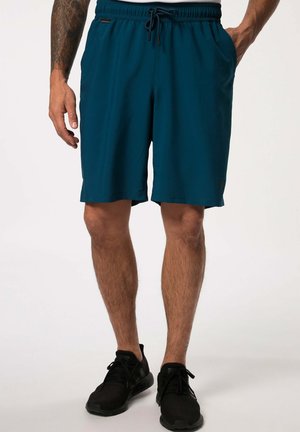 Mies, jolla siniset urheilushortsit, joissa on kiristysnaru, ja mustat lenkkarit, seisoo valkoista taustaa vasten kädet lähellä taskuja.