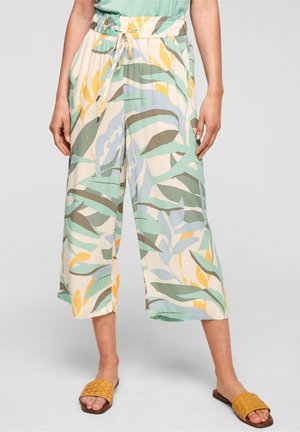 Femme portant un pantalon large à cordon avec un motif de feuilles tropicales vertes, jaunes et bleues et des sandales tressées jaune moutarde.