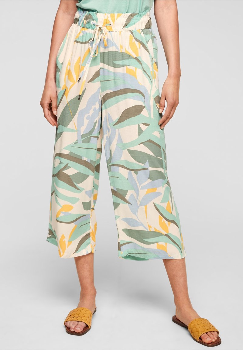 Femme portant un pantalon large à cordon avec un motif de feuilles tropicales vertes, jaunes et bleues et des sandales tressées jaune moutarde.
