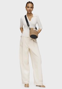 Borsa a tracolla tonda con base nera, etichetta bianca e tracolla regolabile. Indossato un top bianco a maniche corte e pantaloni larghi color crema.