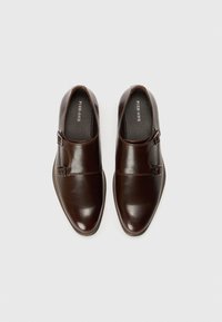 Zapatos de piel marrón con un acabado pulido, que presentan un diseño plano, puntera redondeada y un detalle de hebilla lateral. Forro interior suave en negro.