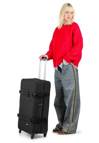 Maleta negra con ruedas y dos correas con hebilla, mango único y acabado liso. Presentada sobre un fondo blanco, junto a unos vaqueros grises de gran tamaño.