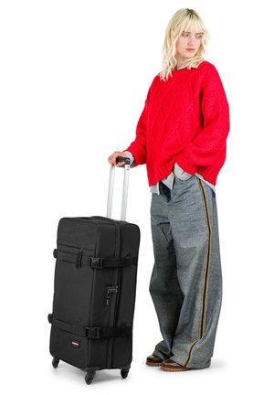 Valigia nera con ruote, due cinghie con fibbia, una maniglia singola e finitura liscia. Posizionata su sfondo bianco, abbinata a jeans grigi oversize.