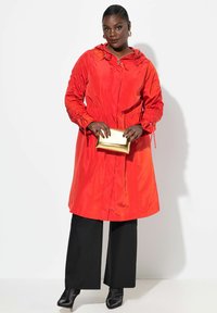 Imperméable rouge à capuche avec des détails de manches bouffantes, associé à un pantalon large noir et des bottines noires. Tenant une pochette dorée.