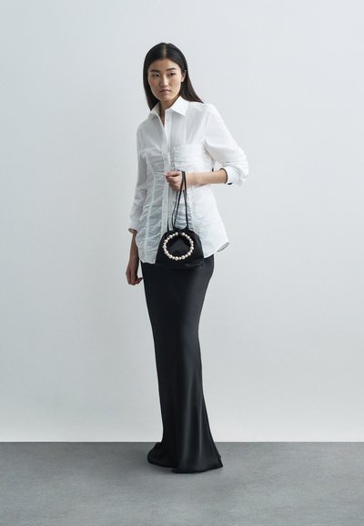 Γυναίκα με λευκό πουκάμισο με ραφή και μαύρη μακριά φούστα κρατά μια μαύρη τσάντα με κυκλικό χερούλι από πέρλες μπροστά σε μια απλή γκρί φόντο.