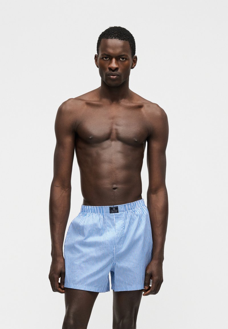 Slanke donkerhuidige man staand, gekleed in lichtblauwe en witte verticaal gestreepte boxershorts tegen een effen lichtgrijze achtergrond.