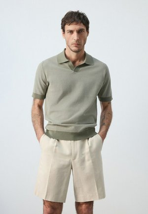 Hombre con cabello corto y oscuro, vistiendo una camiseta tipo polo de manga corta verde claro y pantalones cortos beige hasta la rodilla, de pie con las manos en los bolsillos.