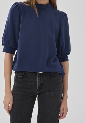 Femme portant un haut bleu marine à manches bouffantes mi-longues et un jean noir taille haute sur un fond uni.