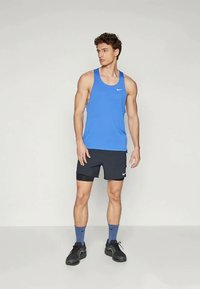 Canottiera blu con scollo rotondo e logo bianco, abbinata a pantaloni corti neri con un dettaglio, indossata con calzini blu e sneakers nere.