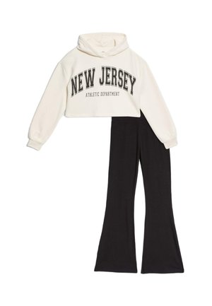 Skrócony biały hoodie z napisem „NEW JERSEY” i czarnymi spodniami typu dzwony. Hoodie ma regulowany kaptur i prążkowane mankiety; spodnie są gładkie.
