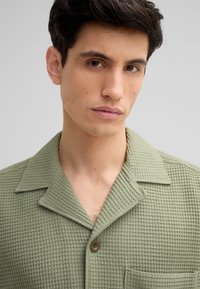 Gestructureerde groene blouse met een kraag, voorzien van een knoop aan de voorkant en een borstzak. Het wafelpatroon zorgt voor een unieke tastkwaliteit.
