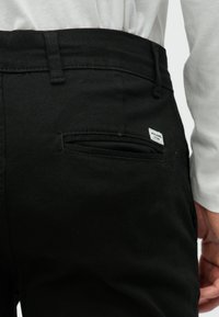 Detail čiernych nohavíc s vreckom na zadnej strane s etiketou, kombinovaných s bielou košeľou s dlhým rukávom, zobrazenie časti ruky osoby.