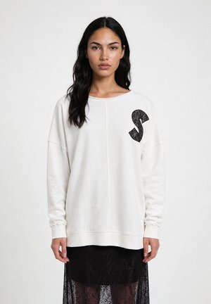 Woman with long dark hair wearing a white oversized sweatshirt with a black lace "S" on the chest and a black lace skirt.