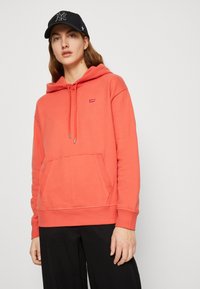 Felpa color corallo con tasca a marsupio, cappuccio con cordino e piccolo logo rosso di Levi's. Il modello indossa pantaloni neri e un cappellino nero.