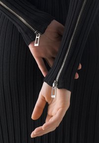 Robe noire côtelée à manches longues avec fermetures éclair argentées le long des bras. Vue en gros plan mettant en valeur les détails des fermetures éclair et la texture.