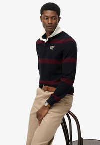 Marineblå og burgunderstribet polo med hvid krave, der har et logo på brystet; parret med beige bukser og et brunt bælte.