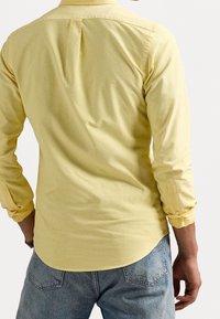 Man i en gul långärmad skjorta med uppvikta ärmar och blå jeans, sedd bakifrån mot en enkel bakgrund.