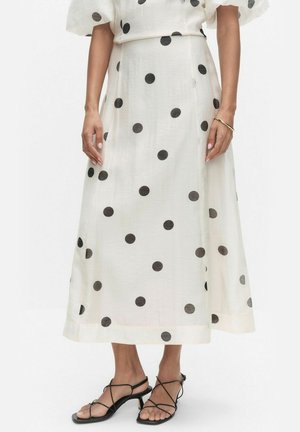 Vrouw draagt een witte midi-rok met grote zwarte polkadots, gecombineerd met zwarte sandalen met lage hak en een gouden armband.