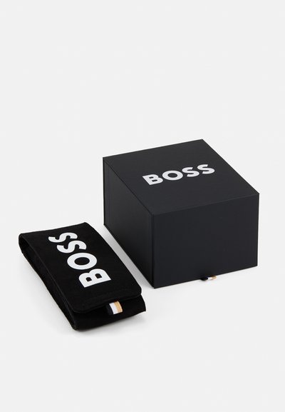 Boîte noire avec logo "BOSS" blanc, accompagnée d'une pochette noire assortie avec logo. La boîte présente une finition lisse et mate ainsi qu'une forme rectangulaire.