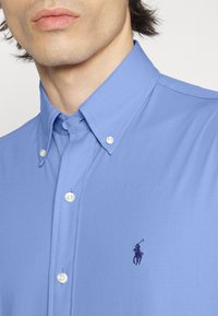 Jasnoniebieska koszula zapinana na guziki z kołnierzem typu spread, białymi guzikami i małym haftowanym logo na lewej piersi. Gładka faktura materiału.